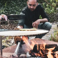 H&ouml;fats TRIPLE 120 Vuurkorf in Cortenstaal met brandende houtblokken, geplaatst op een witte ondergrond.
